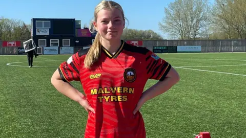 Richard Imogen wearing a red football top standing in a football pitch with her arms on her waist and hair tied back, smiling at the camera.