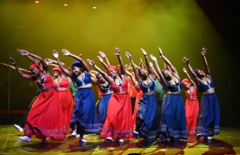 OUPA BOPAPE / GALLO IMAGES / GETTY IMAGES Women dressed in red and blue dance in unison on stage.