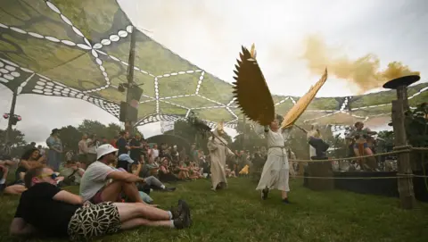 Reuters/JamiJoy A crowd of people sitting on the grass while a ceremony takes place. There are people dressed in white robes with gold wings and masks. There is a fire burning on a pedestal, and a stretched green canvas suspended over everyone.