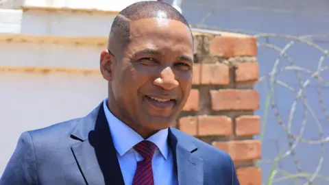 Reuters Duma Boko in a dark blue suit with red and blue stripped tie smiles outside his home in Botswana's capital, Gaborone. Behind him is a brick wall and barbed wire - November 2024