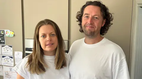 Hannah Gray/BBC A woman with short blonde hair in a white top and a man with brown hair in a curtain style in a white top pose for the camera.