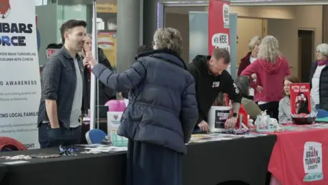 A series of charity stalls, with banners within The Forum, with many people visible in the image on either side of a row of tables, bearing information leaflets and promotional items