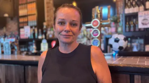Simon Thake/BBC A woman in a black vest and hair tied back stands in front of a bar.