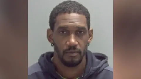 Tyrone Busch has short black hair in his custody photo and is looking at the camera. He is wearing a green top and a dark grey or navy blue hoodie over the top of it.