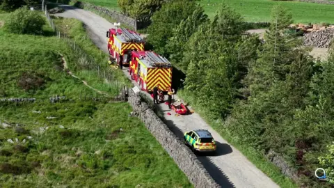 two fire engines and an emergency response vehicle are parked on a country road with fields and tall trees on either side