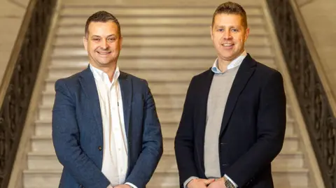 Lidl Two men with short hair smile at the camera. They stand at the bottom of the steps inside Stormont. 