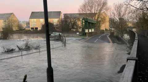 Harriet Heywood/BBC Flood water as taken over a road which can no longer be seen. Towards the back of the image the road re-appears as it goes over a bridge. To the right of the image is a sign for Little Paxton. The sunrise is pink in the sky and there is steam coming off the water in the background.