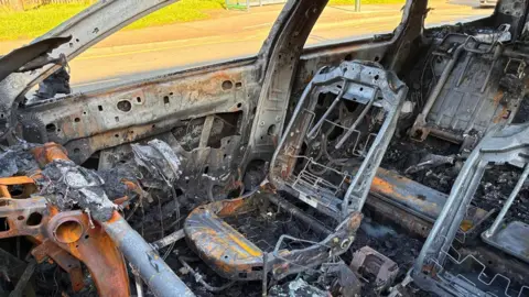The image shows the burnt out shell of a car. All that remains is the metal structure of the seats covered in ash. The windows have been blown out. The steering wheel has melted. 