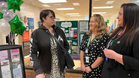 Two social perscribers and a shopper talking outside a shop unit full of NHS posters and leaflets
