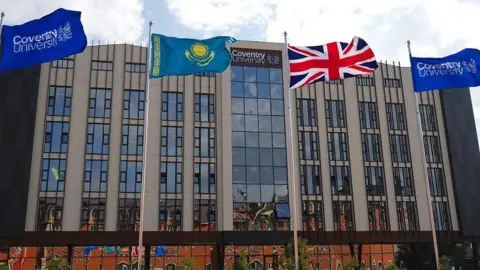 A large grey building with dozens of windows. Two flags with the Coventry University logo are placed either side of a Kazakhstan and Union Jack flag.