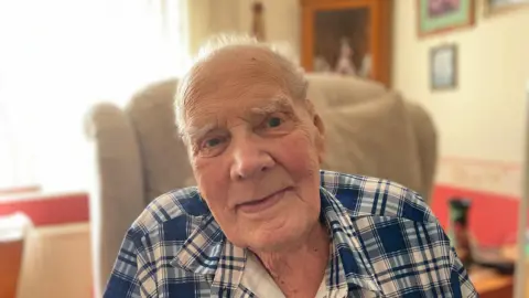 A man is looking at the camera. He has short white hair and he is wearing a blue checked shirt. He is sitting in a high back armchair. There is a glass cabinet behind him and pictures on the wall.