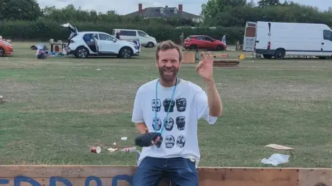 Paul Blakemore, a man with a beard, is carrying a microphone and perching on a wooden sign in a field.
