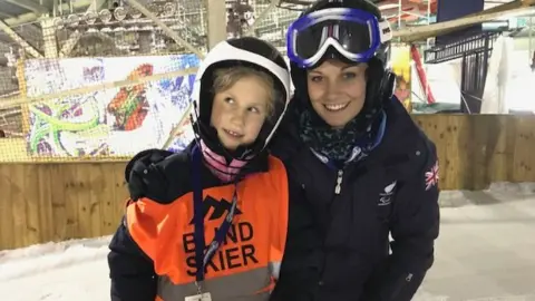 Mendip Activity Centre A close up of Hester and her guiding partner, Ali, who has her arm around her. They are both dressed in ski wear and helmets on an indoor ski slope. Hester has a bright orange tabard on saying 'Blind skier'