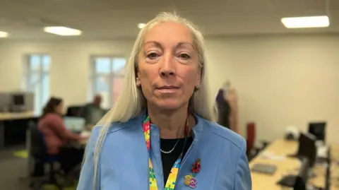 Emma Baugh/BBC News Sarah is wearing a pale blue jacket and a lanyard made up of colourful jigsaw shapes. On her lapel is a red metal poppy. Her hair is long and blonde and is worn down. She is looking directly at the camera. In the background, two people can be seen working at computers.