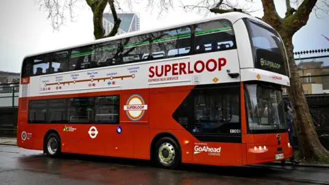 TfL A general view of the new superloop express bus service on a wet floor outside. The bus has superloop written bold in red and a map of the route between Waterloo and Lewisham Centre.