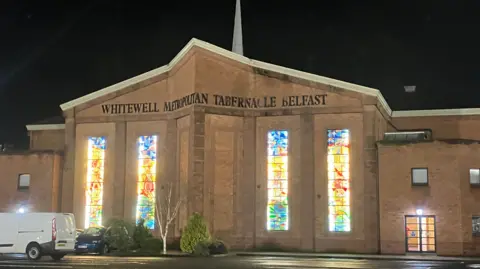 BBC The church is a brown stone building with a white spire coming out of its' roof. There are four large stained glass windows. And a car park in front of the building.