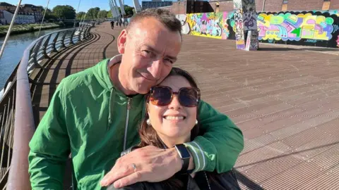 Wayne Ellaway Selfie of a middle aged man and woman smiling, they are on a bridge and it is a sunny day, she is wearing sunglasses he is wearing a green jacket