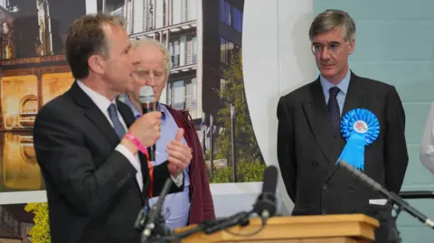 PA Media Jacob Rees Mogg wearing a blue rosette pinned to his suit looking at Dan Norris who is holding a microphone in front of a podium during the general election count result in July 2024