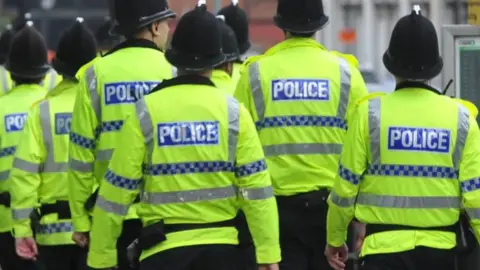 BBC A group of police officers walking away from the camera. The officers are wearing black helmets and yellow high-vis jackets with 'police' written on the back