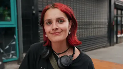 Hannah Karpel/BBC young woman with short red hair in the foreground, wearing a black T-shirt, black over-ear headphones around the neck, a cross-body strap, a nose ring and earrings. The person is facing the camera on a pavement outside a building. Behind the person is a dark tiled wall and metal security shutter, with a turquoise-framed doorway visible on the left and part of a street scene on the right. No bags, bins or markers are visible.