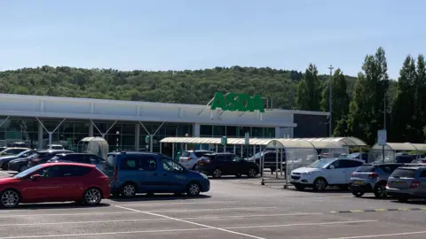 A outside shot of the Asda store in Leckwith. There are a number of cars parked outside with some parked beside trolley bays. 