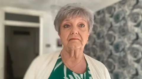 BBC Joanne Horner-Bloomfield, with short grey hair and wearing a white cardigan and green top, looks towards the camera