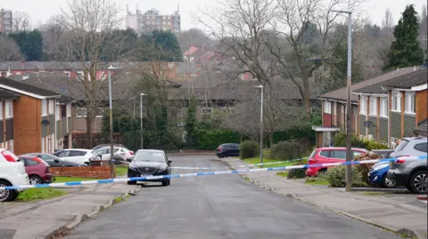 BBC A residential street with cars and houses either side and trees in the background. A police cordon is in place across the middle of the street.