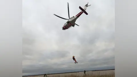 HM Coastguard Southend-on-Sea A person hanging on the end of a cord, from a helicopter, that is flying in the sky. The helicopter is white and red, there is a large open sky and then reeds below. 