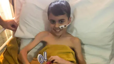 A young boy who is very skinny has a breathing tube in his nose and is sitting a hospital bed covered with a yellow blanket and candles in the shape of a ten