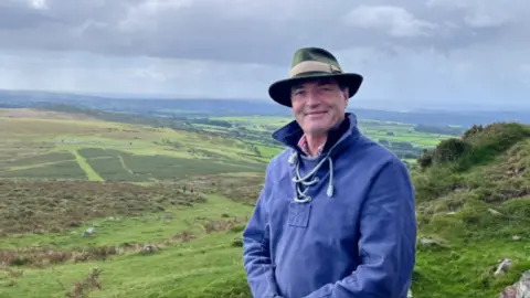 David Braine standing to the right of the frame with a rolling green landscape behind. He is wearing a blue top and a wide-brimmed hat. He is looking at the camera and smiling.