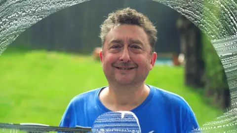 Madoc Roberts Steven pictured cleaning a window, seen from the inside of the window. There are soap marks around the edges and a clear glass in front of his face. He is smiling and wears a blue t shirt. Green lawn behind him. 