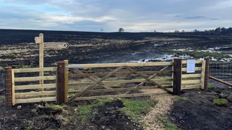 Mark Young/North York Moors National Park Authority A brand new wooden gate and signpost are situated in a bleak landscape of burned, blackened  vegetation.  The area behind leading to the horizon is wet and muddy.  The sky is partially overcast with a small patch of blue peeking through.