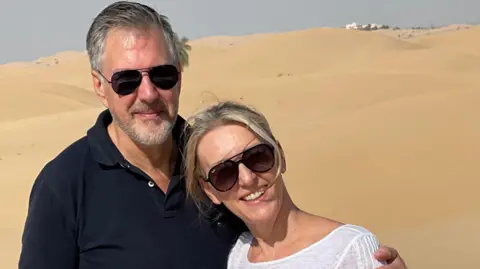 Angela Hudson Angela Hudson, in a white top and aviator sunglasses, and her husband Chris, in a black polo shirt, with sand dunes behind them.