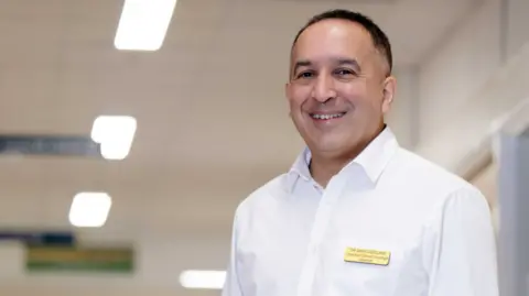 Gloucestershire Hospitals NHS Trust Sam is in a mid-shot, standing in a hospital environment which is blurred in the background. He is wearing a white shirt and a badge with his name and title on.