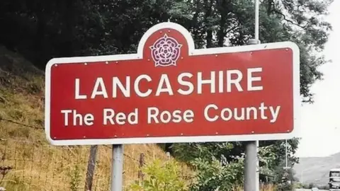 ANTBEX74 A sign for Lancashire featuring the county's red rose symbol, and the words 'The Red Rose County' underneath. Trees and a grass verge can be seen behind it.