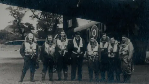 Stuart Green Eight RAF airmen stand in a line in front of their Stirling bomber. They are dressed in full flight suits.