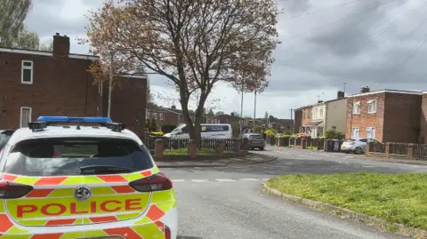 A police car is parked outside properties in Willis Pearson Avenue. A grass verge can be seen on the right with apartments/small blocks of flats visible off to the right and left of the image. A large stands outside one block.
