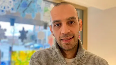 A man with a shaved head and grey jumper looks at the camera. Behind him a blue and yellow window display is blurred.