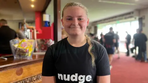 Maisie is wearing a black Nudge and Suntera-branded T-shirt and sits beside a wooden bar inside a clubhouse. The background shows several people gathered in a large room with red carpeting, bright windows, and a television mounted on the wall.