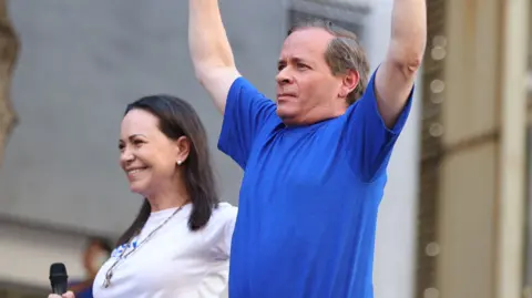Getty Images Juan Pablo Guanipa (right) holds his arms in the air while wearing a blue t-shirt beside Maria Corina Machado (left), wearing a white t-shirt and holding a microphone.