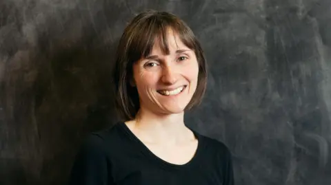 Cat Lane Elaine Midgley, standing in front of a black wall, she is smiling and has shoulder length dark hair. She is wearing a black top. 