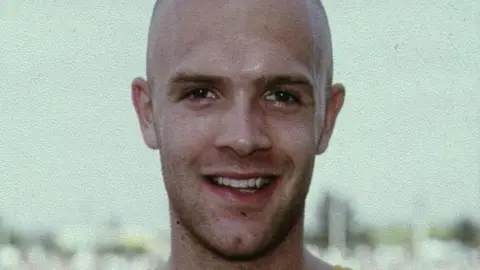 Curtis Robb in a photograph from earlier in his career. He has a shaved head and is smiling for the camera.