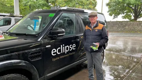 BBC/Julia Lewis A man wearing a flat cap, and blue and beige bodywarmer over a blue jumper, stands next to a black 4x4 vehicle advertising his equine veterinary practice, on a petrol station forecourt.