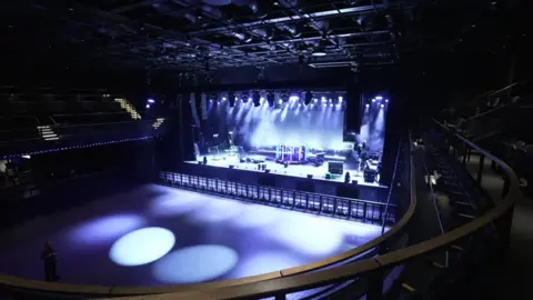 Empty concert venue with stage lighting, instruments set up, and seating around the auditorium. The lighting has a blueish tint.
