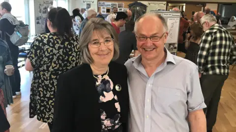 Janice Ross Cindy and David Brockington smile at the camera. Cindy's straight grey hair reaches her shoulders. She wears glasses, a black jacket and a pink-and-black top. David has short white hair, glasses and wears a grey, short-sleeved shirt.