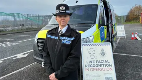 ACC Jennifer Wilson, in her black police uniform, stands in front of a marked police van next to an A-Board sign reading 'Live Facial Recognition in Operation'