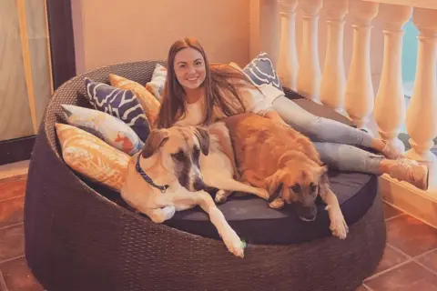 Caitlyn Chalmers A woman smiles while lying on a round outdoor wicker daybed with two large dogs. The daybed is covered with patterned cushions and sits on a tiled balcony with white balustrades. Warm lighting suggests evening, and the sea is visible in the background.