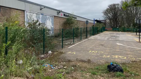 LDRS A sign reading 'Wakefield Westgate' stands atop a brick wall. A fenced off and apparently empty car park stands adjacent. Weeds and plants can also be seen.