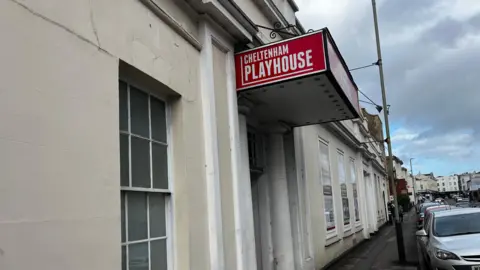 External shot of Cheltenham Playhouse, pictured from the side showing its red porch sign. The building is white.