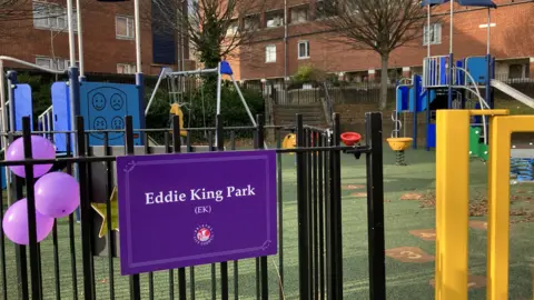 BBC A close up shot of the black metal railings around the perimeter of the play park. Attached to the railings is a rectangular purple sign which says 'Eddie King Park' along with the Bristol City Council logo. Beside the sign there are three purple balloons tied to the railings. Inside the park you can see a swing set, climbing frames, see-saw and slide.